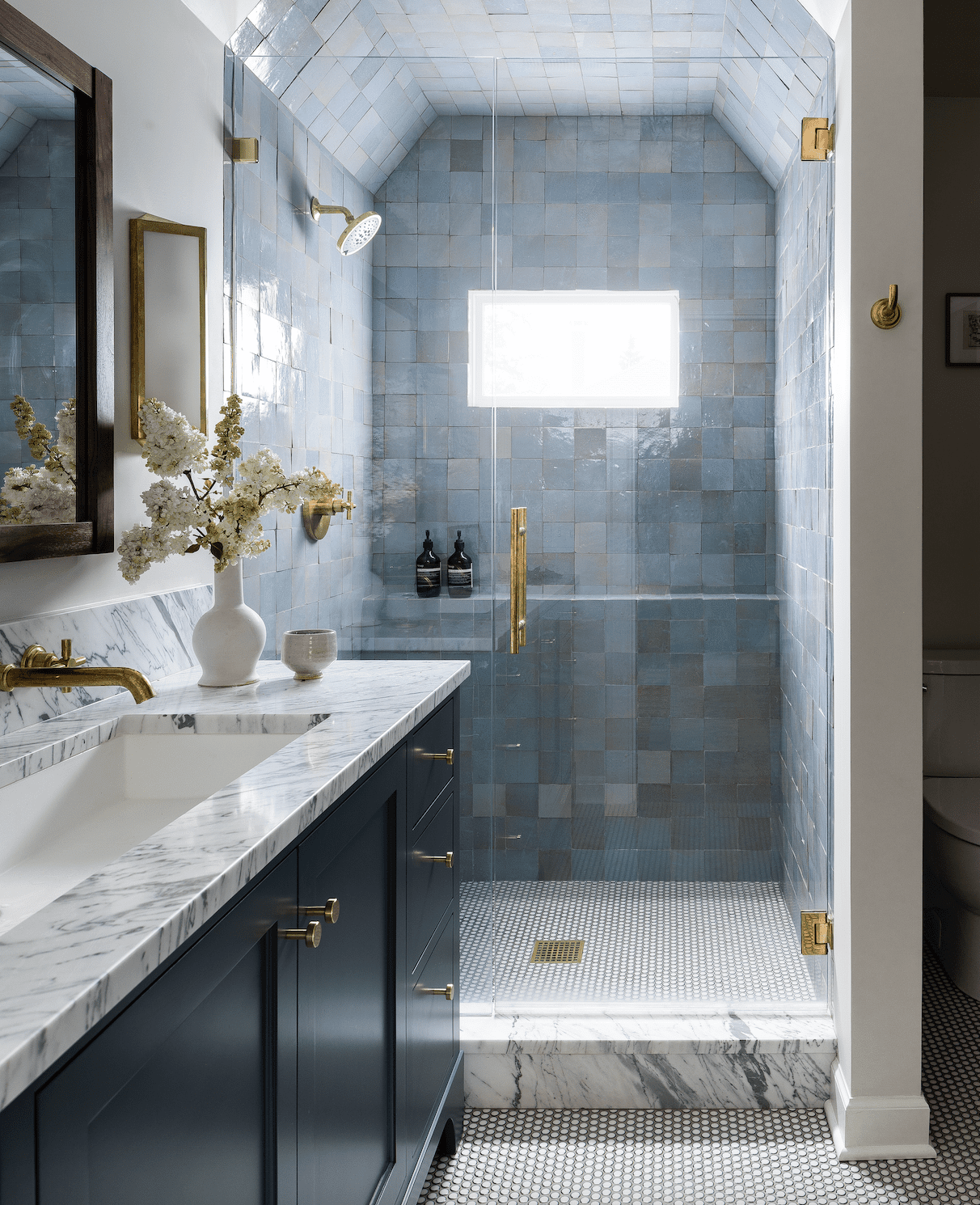 Walk-in shower with blue zellige tile, brass fixtures, frameless glass door and marble curb